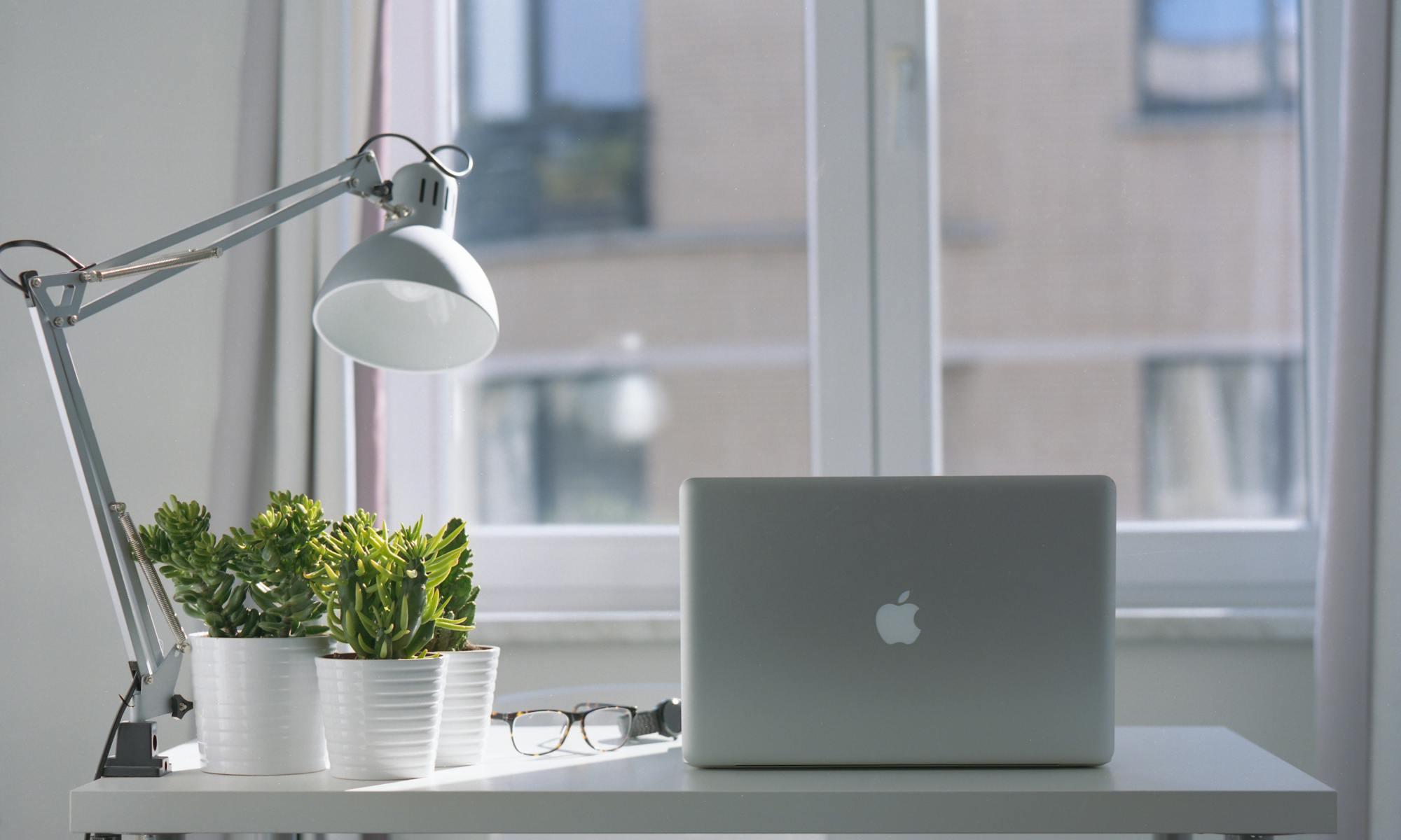 Window in back ground, blurred image of offices outside. Modern desk in front of window, with angle poised lamp in grey to the left with x3 small potted green plants. glasses on desk and open Apple laptop in the centre. Image suggestive of contemporary simple but effective business.