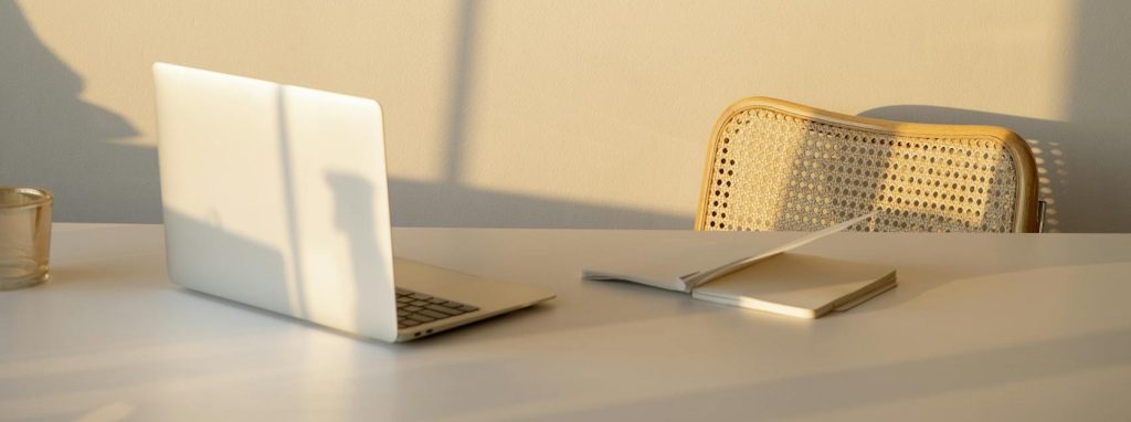 Gentle lighting. desk with open silver laptop and notebook with cain chair behind the desk. Image taken from across the desk. Shadow of sun coming through window reflected on laptop and chair. This image is used to suggest that Therapeutic Consultancy is a contemporary business, slick and professional in it's approach