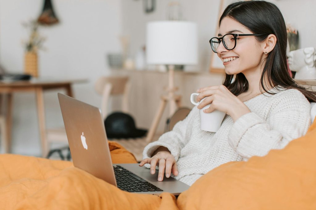 Female mid to late 20's sat on sofa, wearing glasses and smiling. Holding a cup of coffee to her chest and working on a silver Apple laptop with the other hand. This image is used to represent the demographic that Therapeutic Consultancy is designed to attract